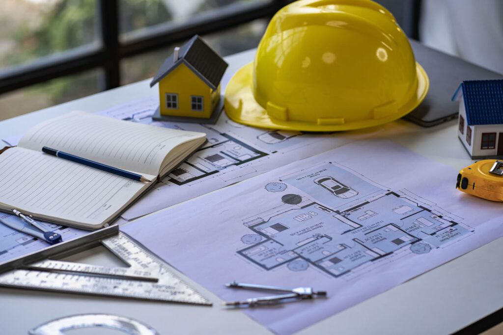 Architectural plans on a table with a yellow hard hat, scale model houses, notebook, pen, measuring tape, and drafting tools.