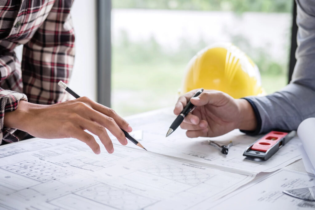 Two individuals discuss architectural blueprints on a desk with a yellow hard hat, level, and pencils.