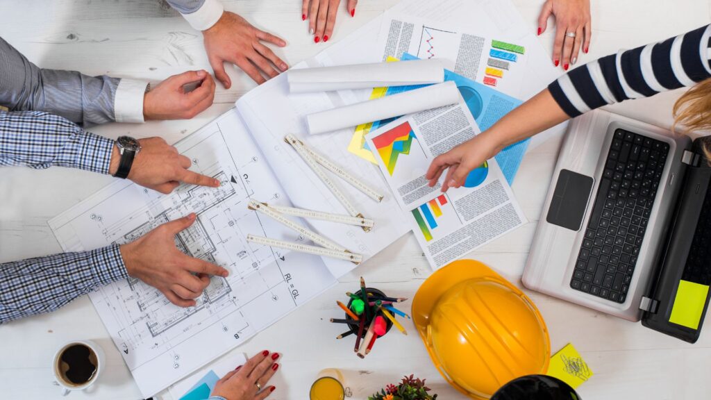 Construction professionals reviewing blueprints and performance charts on a desk with laptops and safety helmets