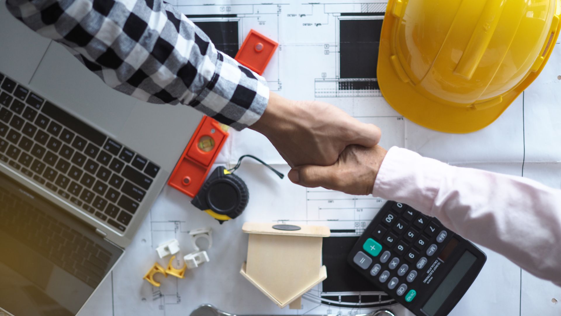 Contractors shaking hands over construction blueprints with a calculator, laptop, and hard hat, symbolizing planning and financial strategy.