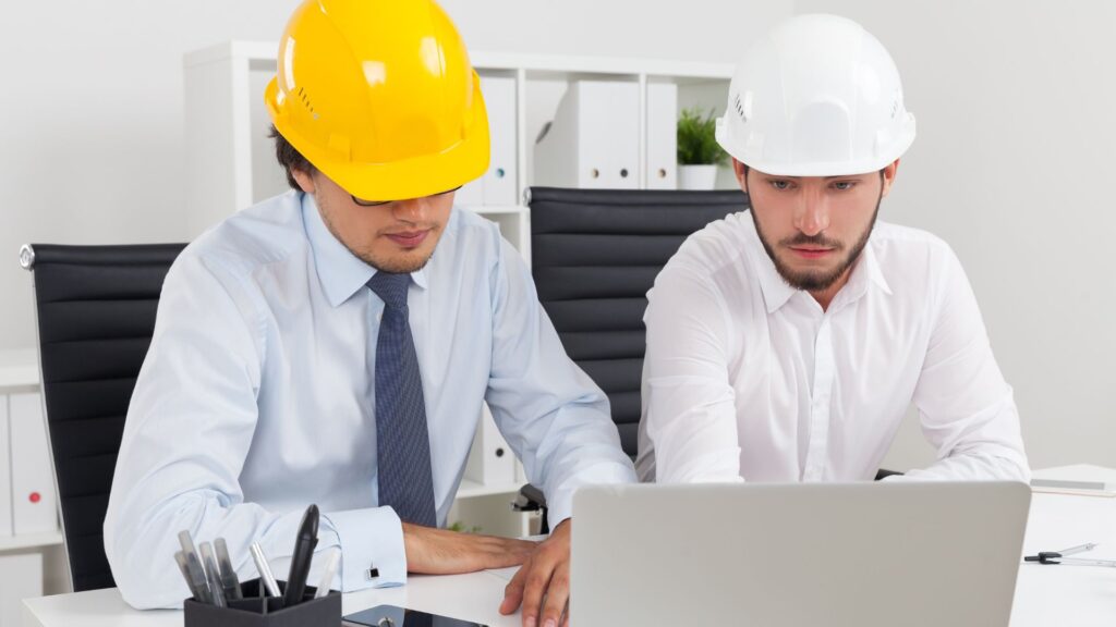 Two construction engineers wearing safety helmets analyzing KPI data on a laptop in an office